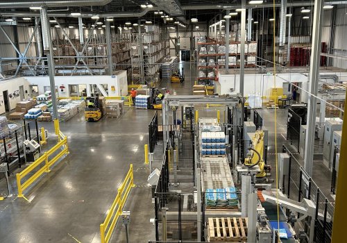 Grande installation industrielle avec des convoyeurs automatisés, des palettes de produits emballés, des chariots élévateurs en circulation et des rangées d’étagères de stockage dans un vaste entrepôt.