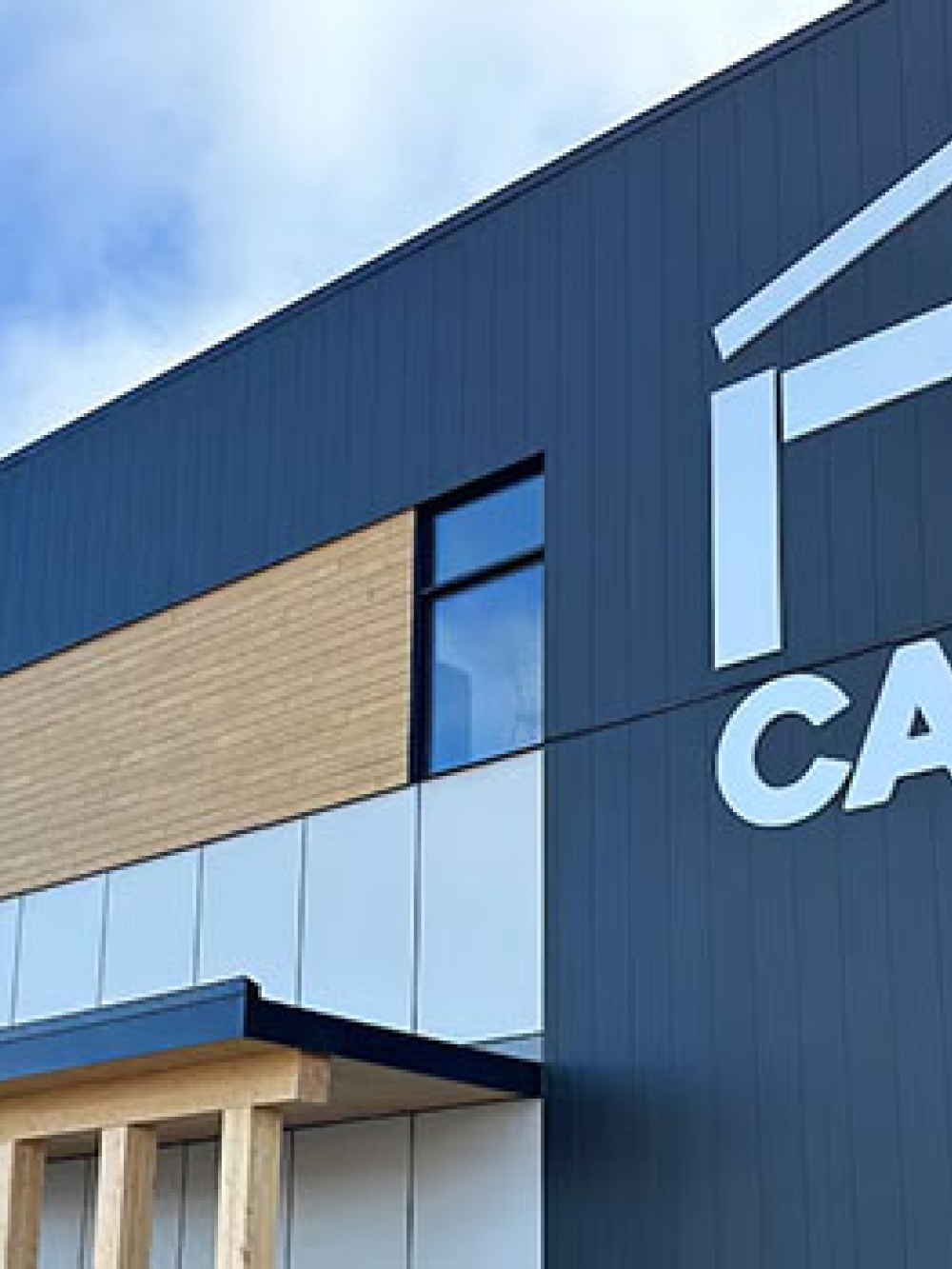 Façade moderne d’un bâtiment industriel gris et bois portant le logo « NT CAMPS », sous un ciel bleu.