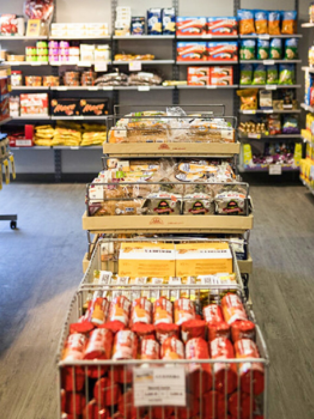Rayon d’épicerie avec des étagères remplies de produits alimentaires et ménagers, incluant des conserves, des bouteilles, des boîtes et des paniers de produits au centre de l’allée.