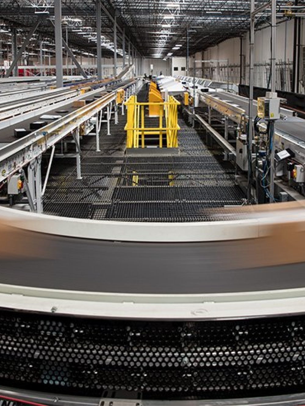 Système automatisé de convoyeurs industriels transportant des colis dans un grand entrepôt logistique équipé de structures métalliques et de mécanismes de tri.