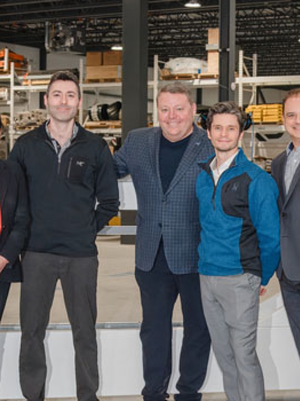 Groupe de sept personnes debout à l’intérieur d’une usine, posant devant une grande structure métallique avec de l’équipement industriel en arrière‑plan.