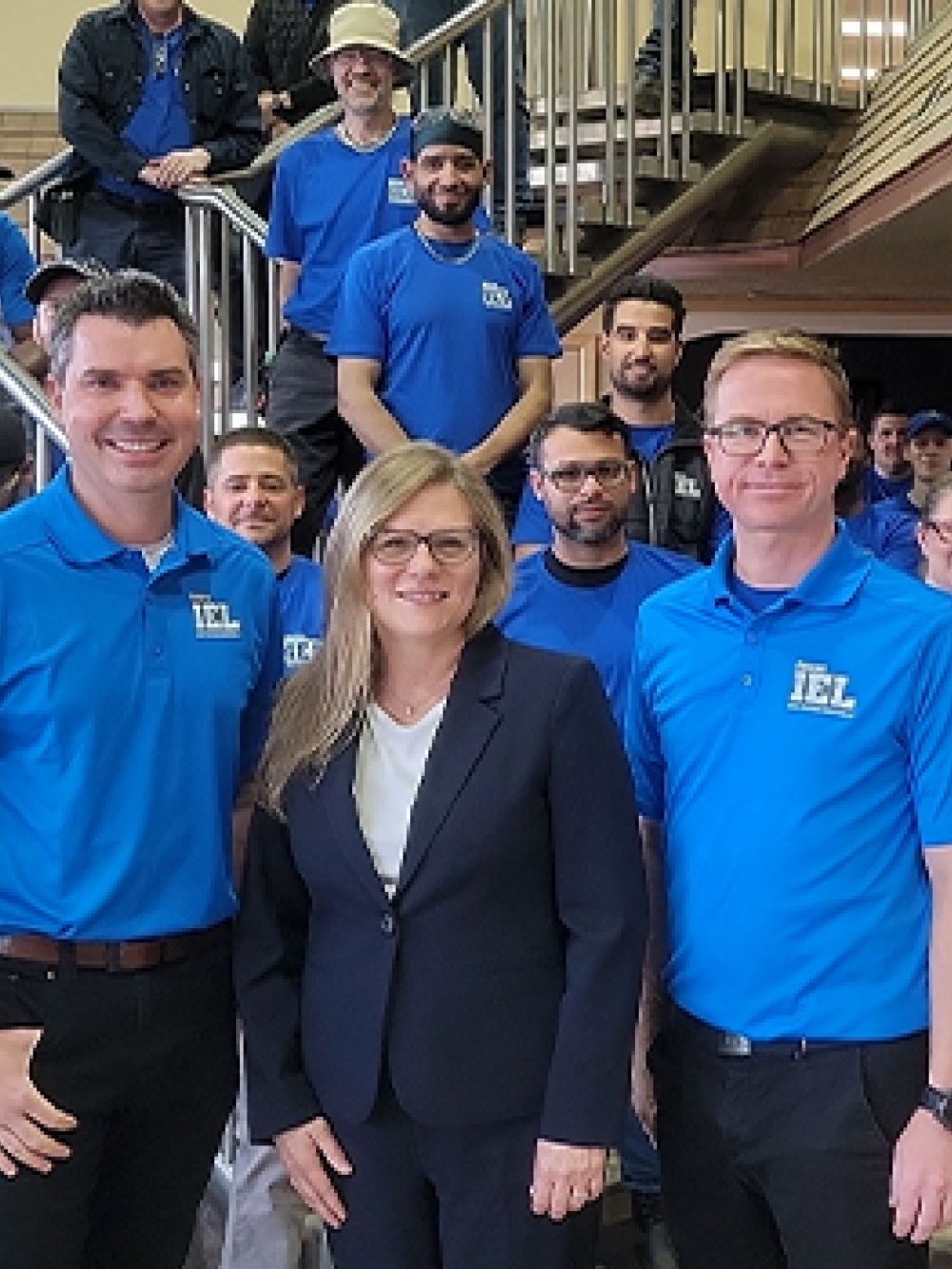 Groupe de personnes réunies dans un hall intérieur, plusieurs portant des chandails bleus avec le logo IEL, posant devant un escalier métallique dans un environnement industriel.