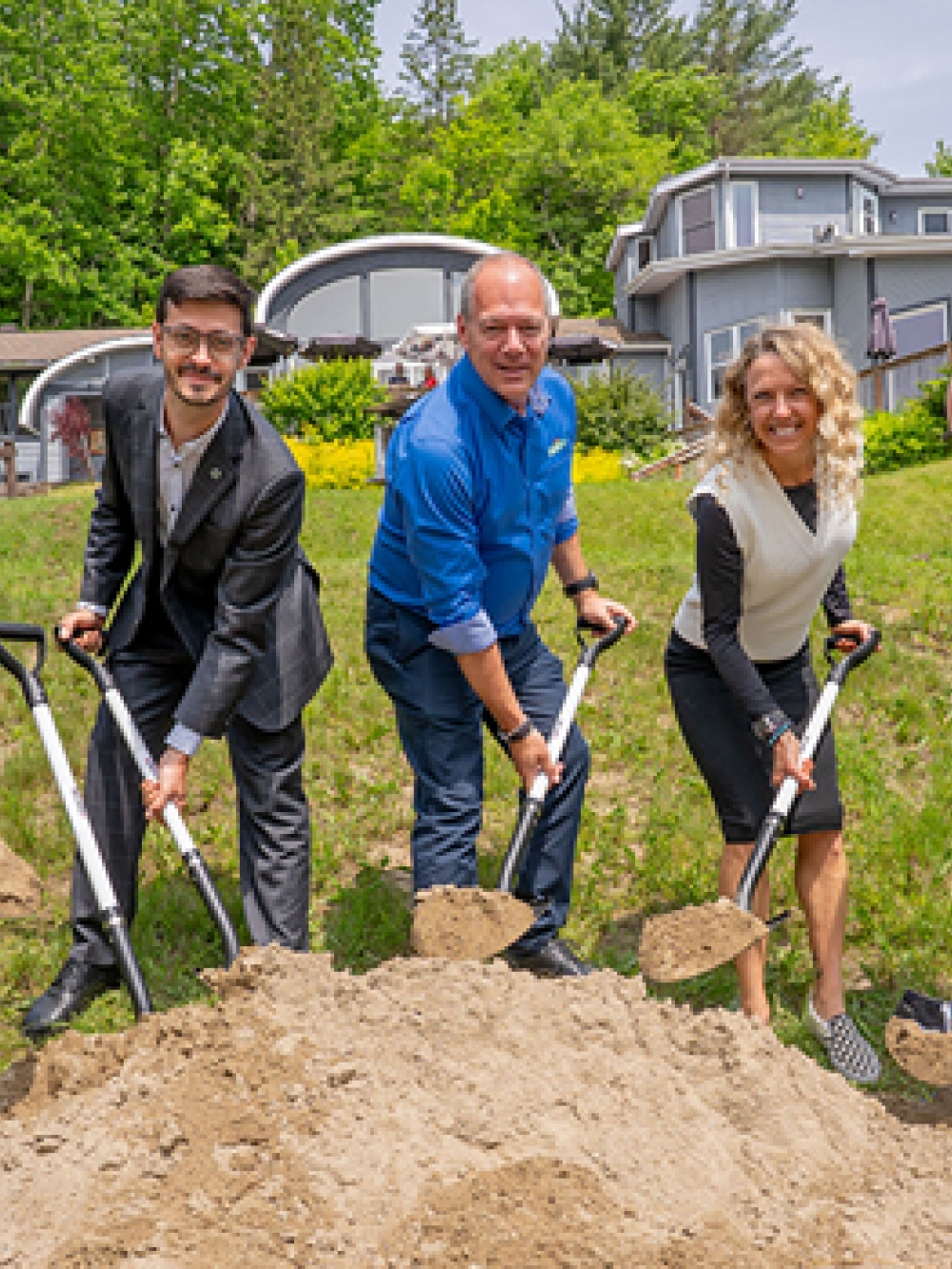 Groupe de personnes participant à une pelletée de terre symbolique sur un terrain gazonné, chacun tenant une pelle remplie de sable, avec des bâtiments et des arbres en arrière‑plan.
