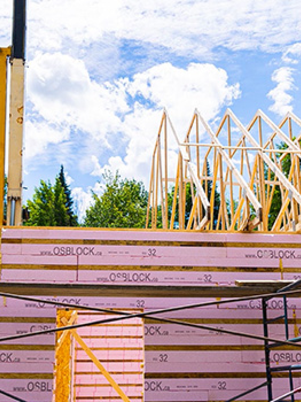 Structure de bâtiment en construction avec des panneaux isolants OSBLOCK et une charpente en bois, accompagnée d’un camion et de machinerie sur un chantier entouré d’arbres.
