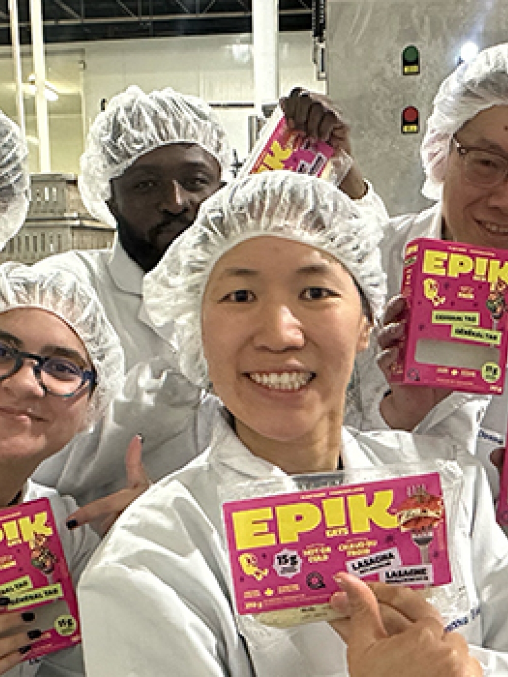 Groupe de personnes en tenue de production alimentaire tenant des emballages de produits EPIK dans une usine, devant un équipement de transformation.
