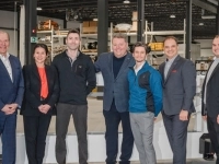 Groupe de sept personnes debout à l’intérieur d’une usine, posant devant une grande structure métallique avec de l’équipement industriel en arrière‑plan.