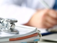 Stéthoscope posé sur une pile de documents médicaux, avec une personne en arrière‑plan écrivant à un bureau.