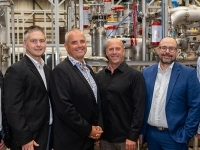Groupe de personnes debout dans une usine, devant un ensemble de tuyaux, de valves et d’équipements industriels en acier inoxydable.