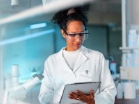 Personne en sarrau de laboratoire tenant une tablette électronique dans un environnement scientifique moderne, entourée d’équipements et de verrerie.