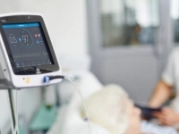 Appareil médical affichant des données de surveillance posé près d’un patient alité, avec un professionnel de la santé en arrière‑plan.