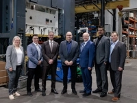 Groupe de personnes debout dans une usine, posant devant un grand équipement industriel entouré de matériel de production et de rayonnages métalliques.