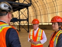 Trois personnes portant des casques et des vestes de sécurité orange discutent à l’intérieur d’un grand bâtiment industriel, près de structures métalliques et d’équipements.