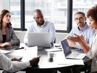 Réunion de travail autour d’une table, où plusieurs personnes échangent et utilisent des ordinateurs portables dans une salle de conférence lumineuse.