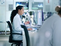 Des personnes en blouse de laboratoire travaillent sur des ordinateurs et des microscopes dans un environnement de recherche moderne, entouré d’écrans d’analyse et d’équipements scientifiques. 
