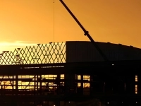 Structure métallique d’un bâtiment en construction, silhouettée devant un ciel orange au coucher du soleil, avec une grue visible au-dessus du chantier.